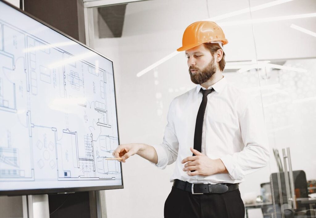 Architect in hard hat reviewing blueprints on a screen, discussing plans in modern office.